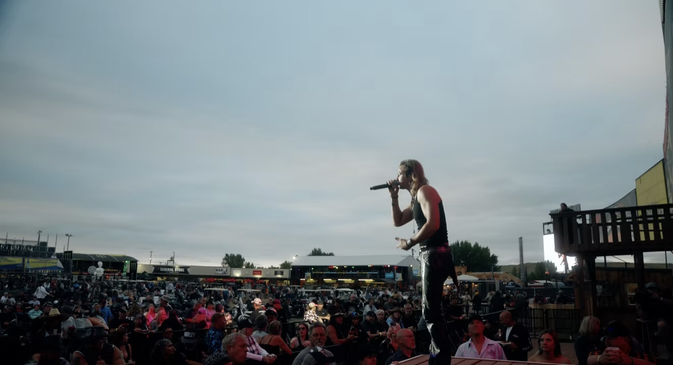 Performer on stage with microphone addressing large crowd outdoors under cloudy sky