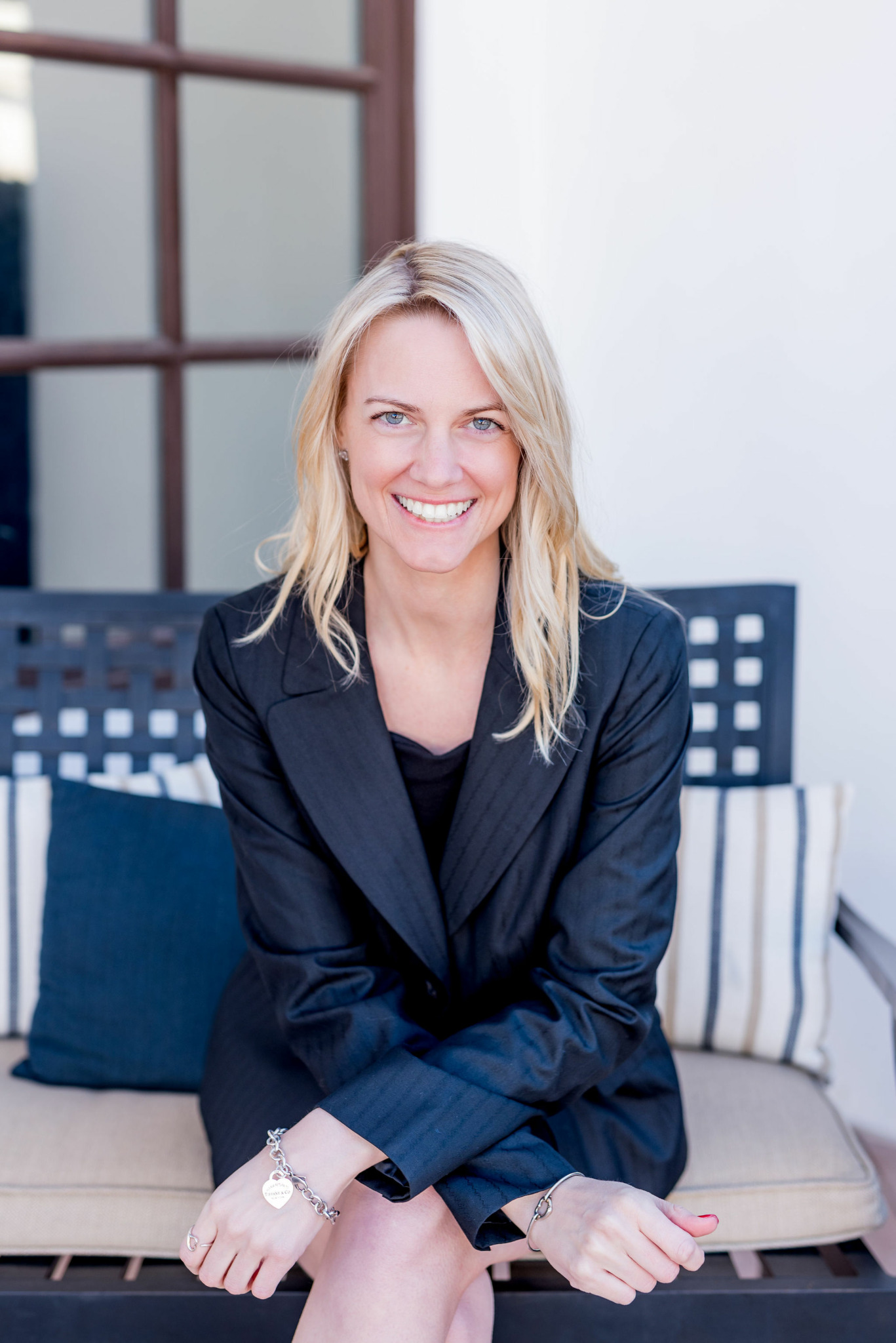 Smiling woman with blonde hair in a dark blazer sitting on a sofa with cushions, in a bright room.