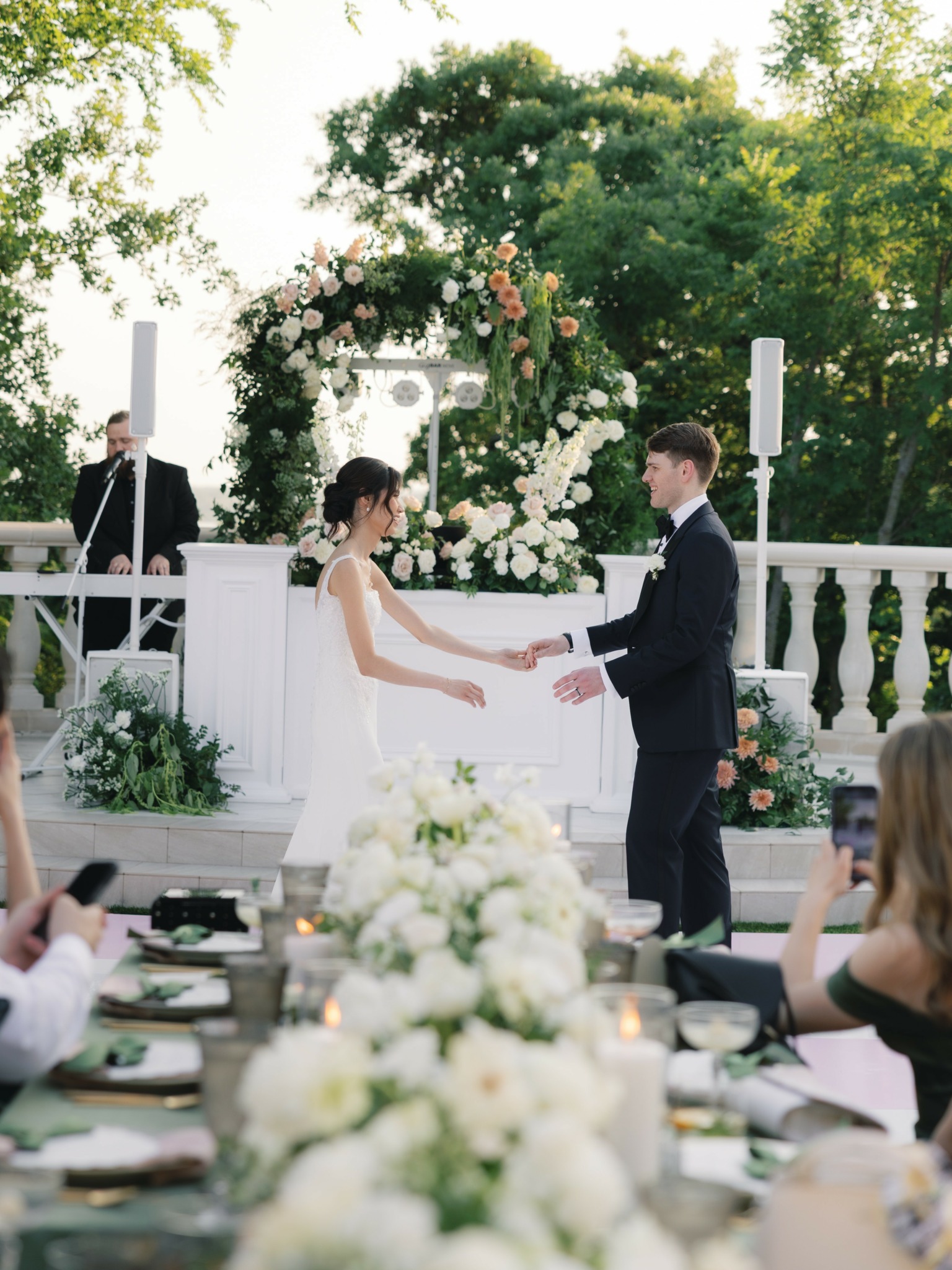 Bride and groom holding hands during outdoor wedding ceremony with floral arch and guests seated at tables.
