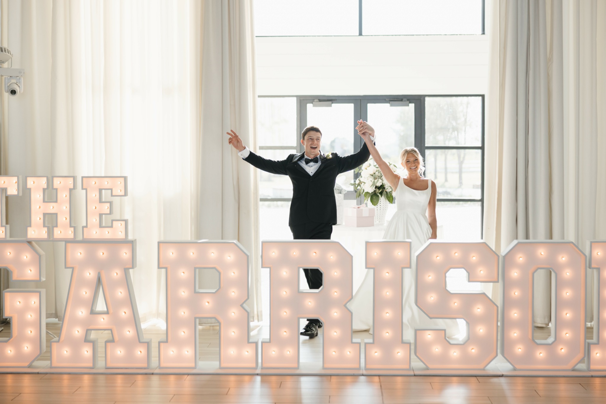 A bride and groom stand behind large illuminated letters spelling 'GARRISON' in a bright room with large windows and curtains.