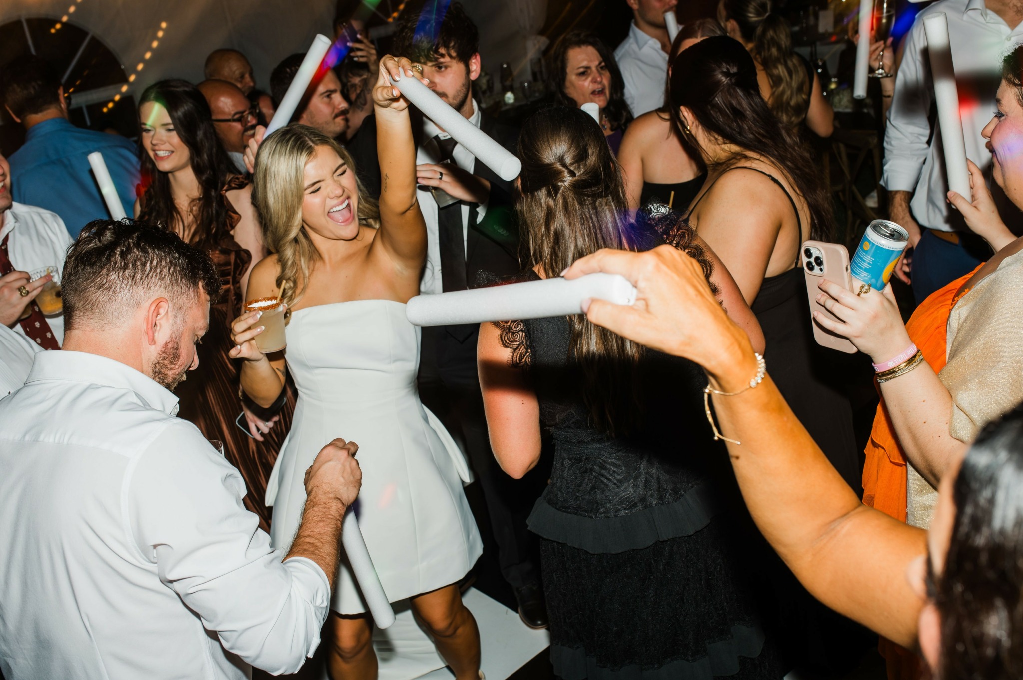 People dancing and celebrating at a party, some holding glow sticks and drinks, with a lively atmosphere.