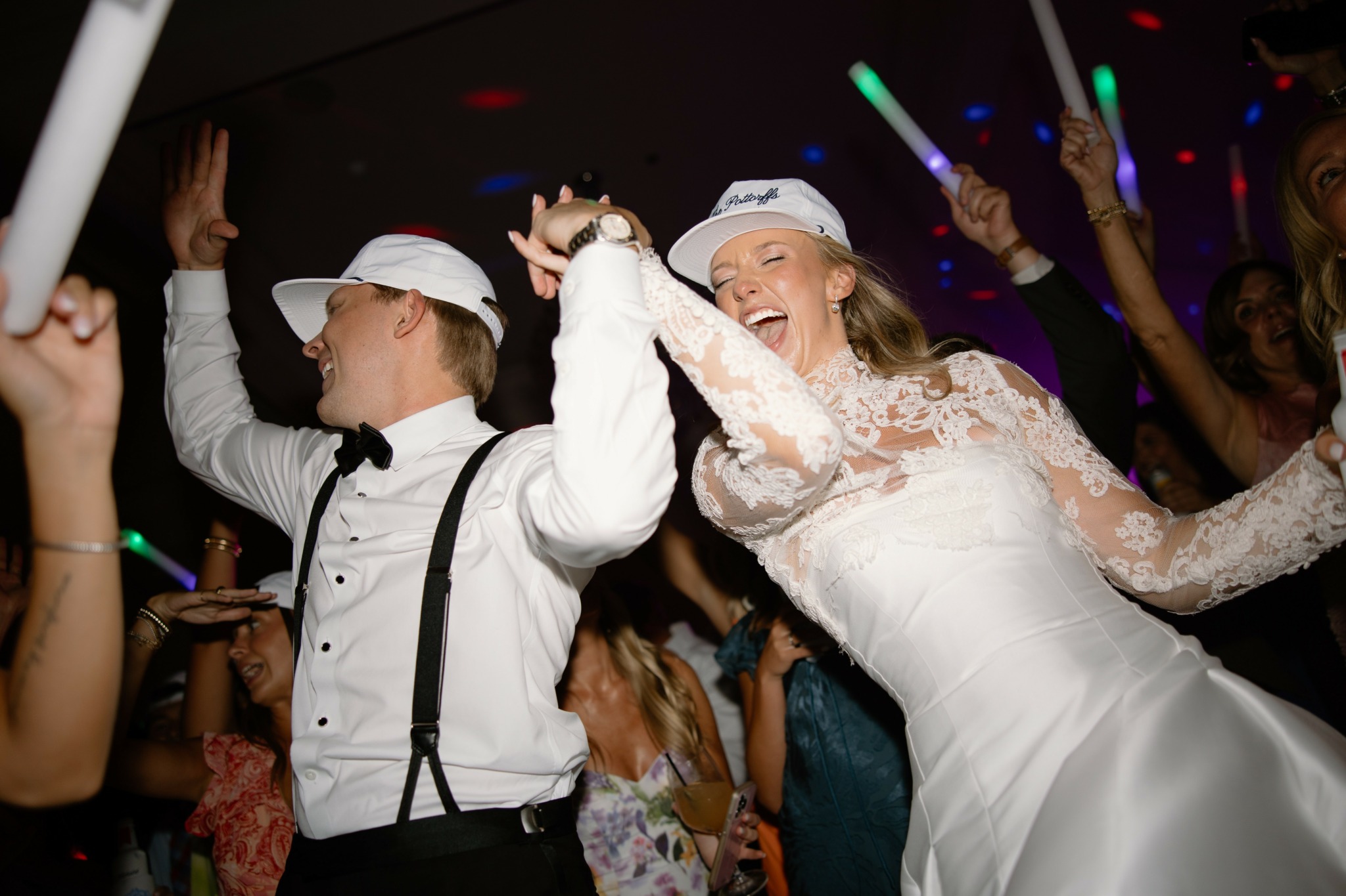 People dancing at a celebration, woman in white dress smiling, others raising hands, colorful lights in background.