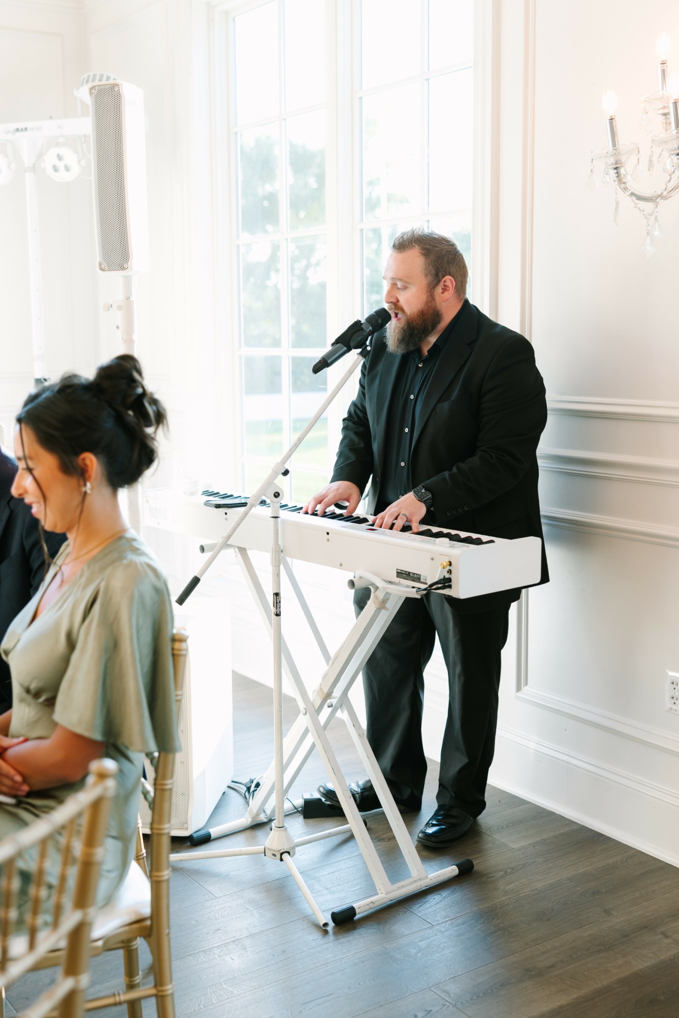 Man singing into microphone while playing keyboard at a bright indoor event, with a woman seated nearby.
