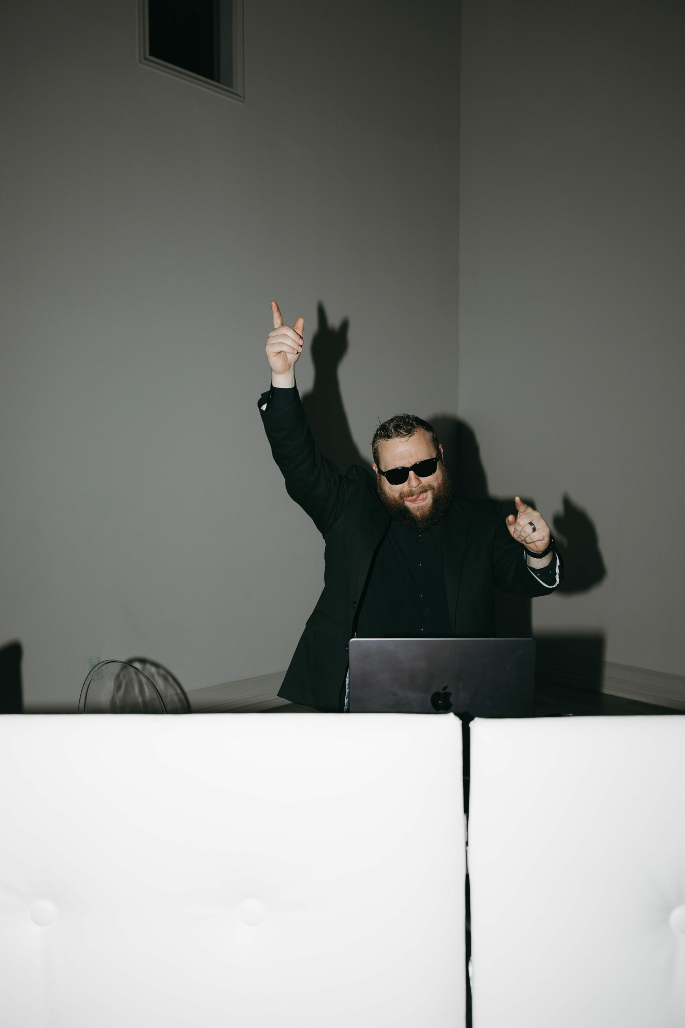 Person wearing sunglasses and black jacket raising one hand with two fingers up, standing behind a white table.