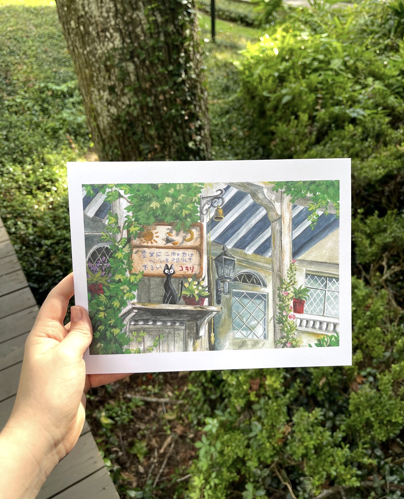 Hand holding a colorful illustration of a house with trees and plants outside, with a garden and a tree in the background.