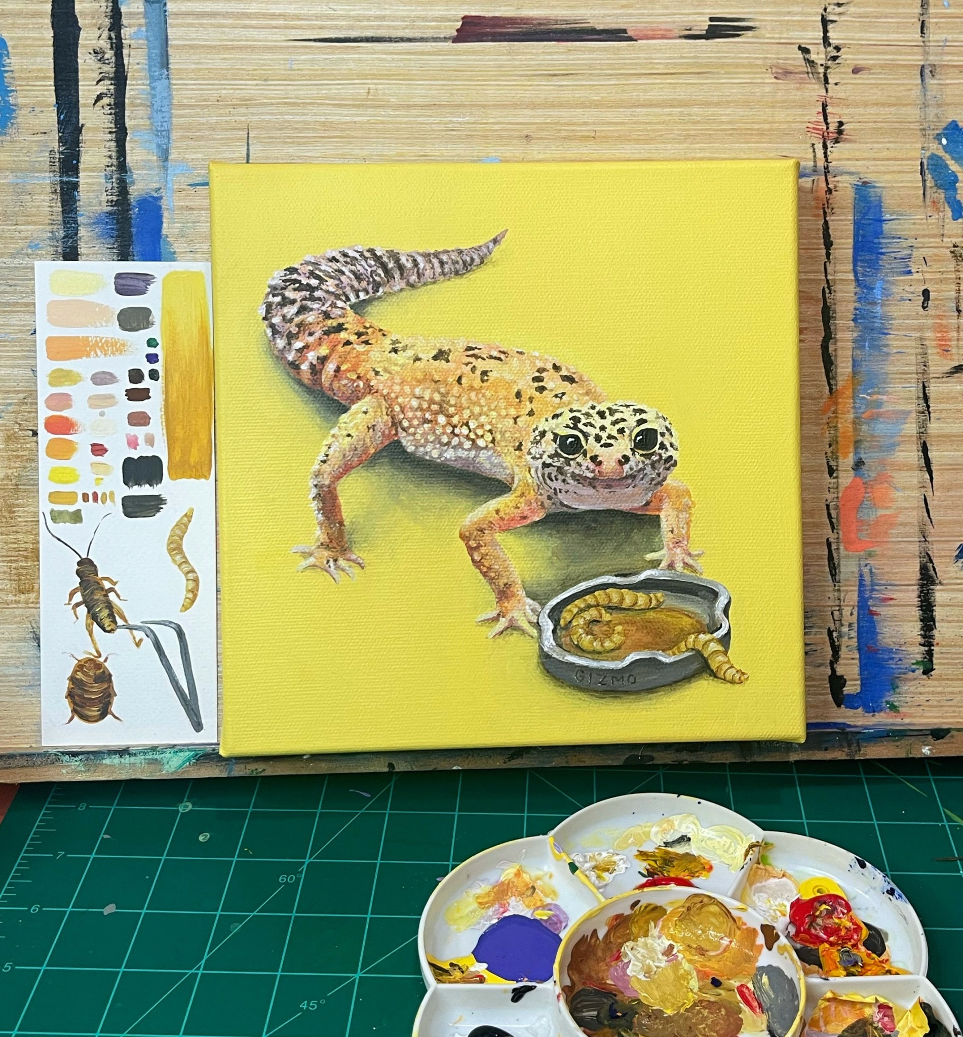 Painting of a gecko with a yellow background, next to a small dish with a snake inside, on a green cutting mat.
