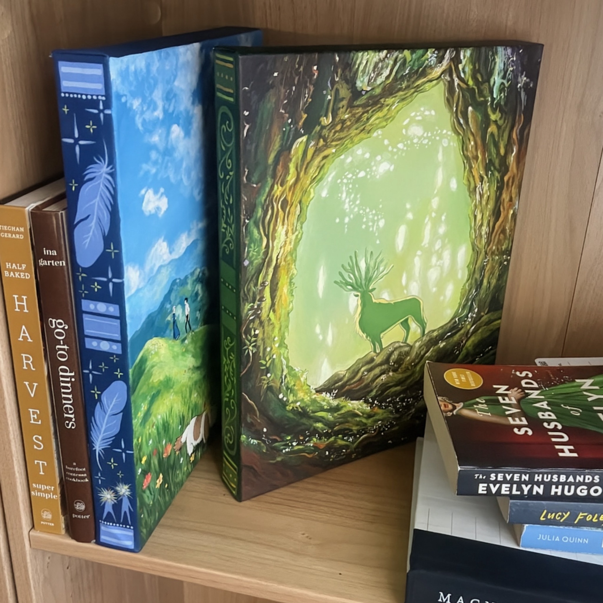 Children's books and a large illustrated book with a deer in a forest scene on a wooden shelf.