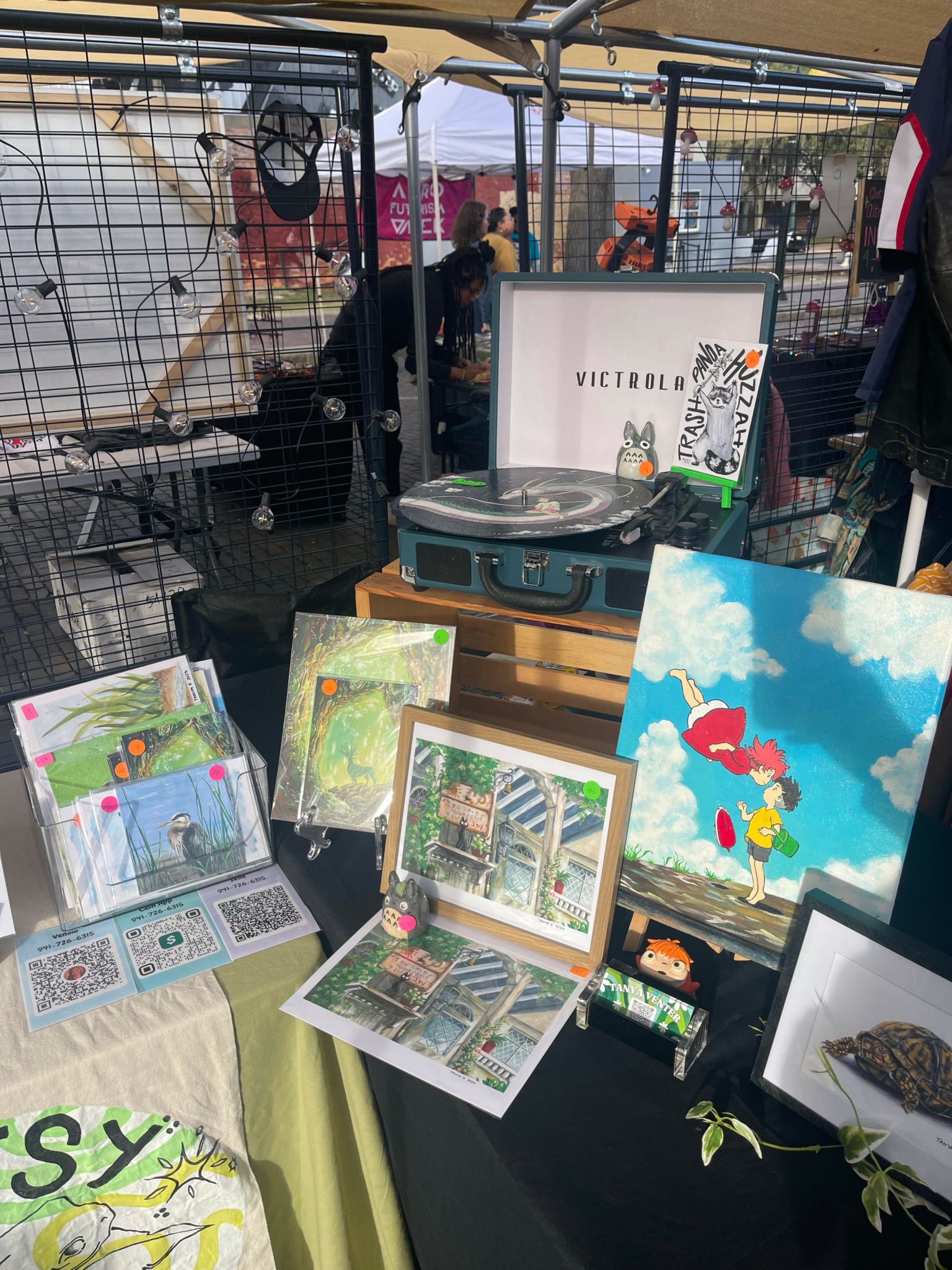 Market stall displaying artwork, framed pictures, and a record player, with people and tents in the background.