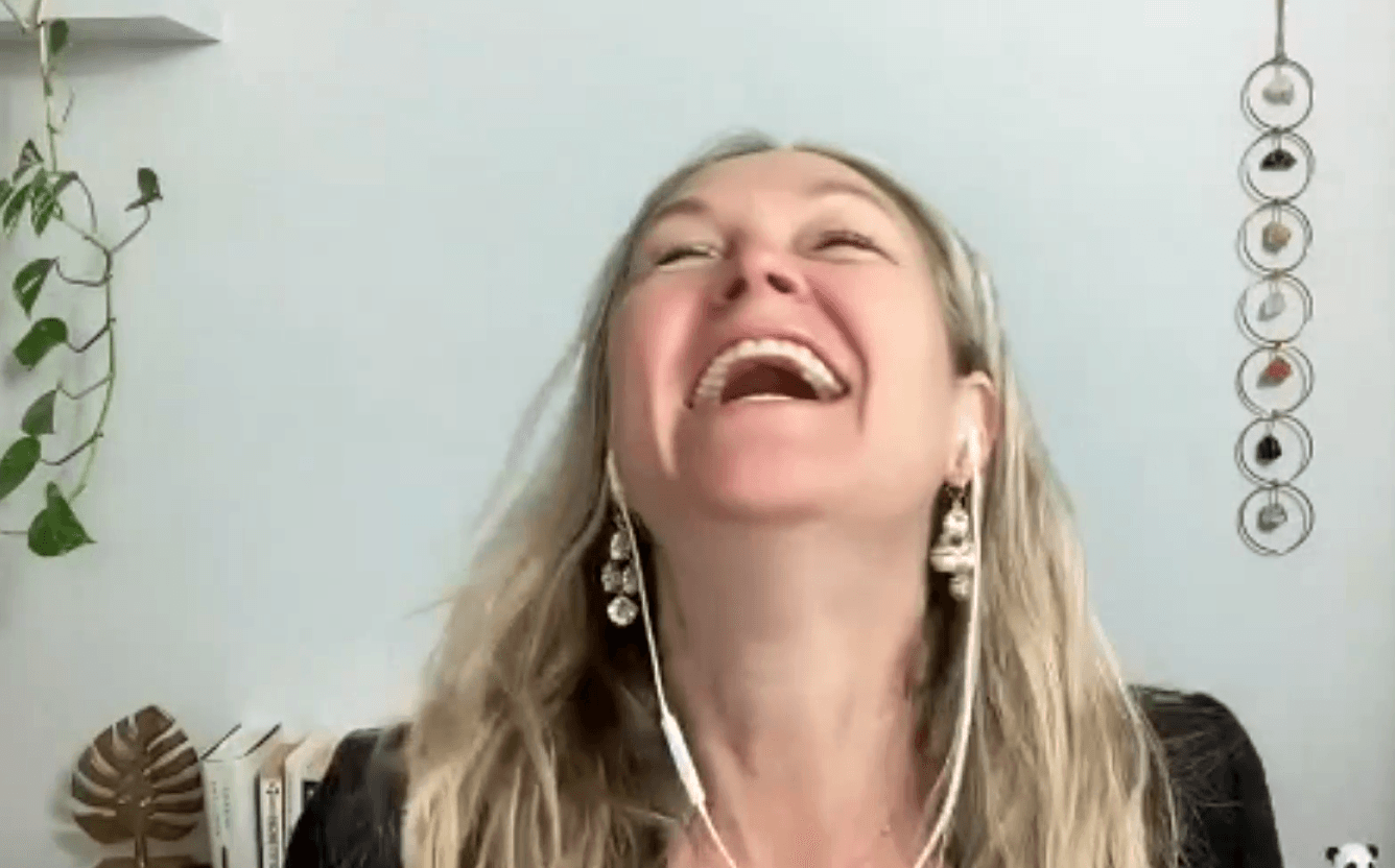 Woman with long blonde hair laughing, wearing earrings, in a room with a plant and decorative hanging objects