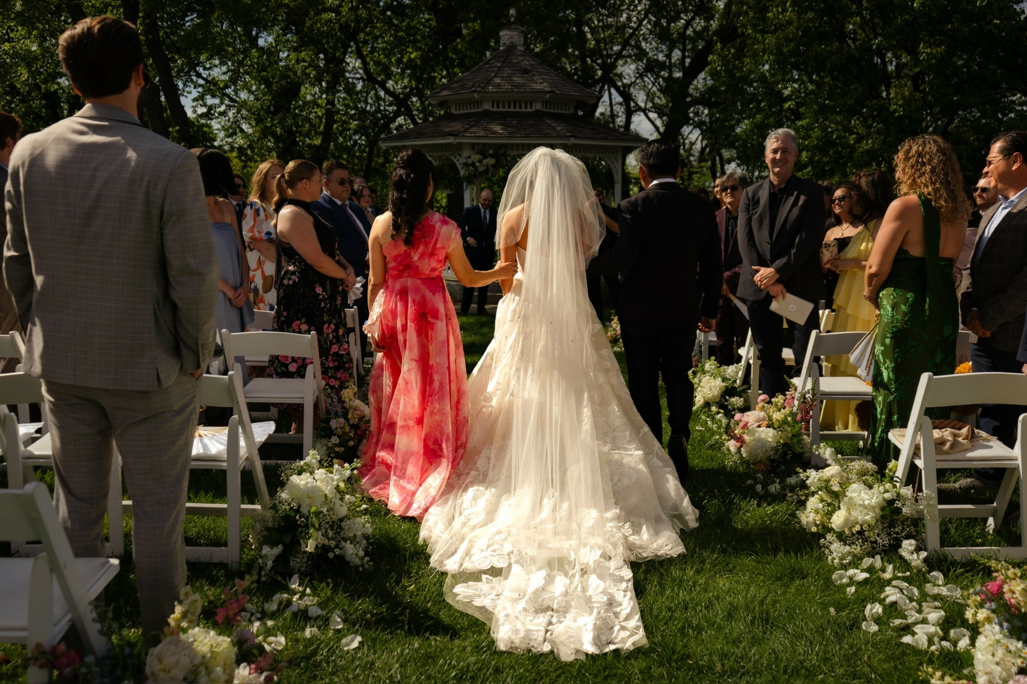 Bride in a white wedding gown walking down an outdoor aisle with guests on both sides, floral decorations, and trees in background.