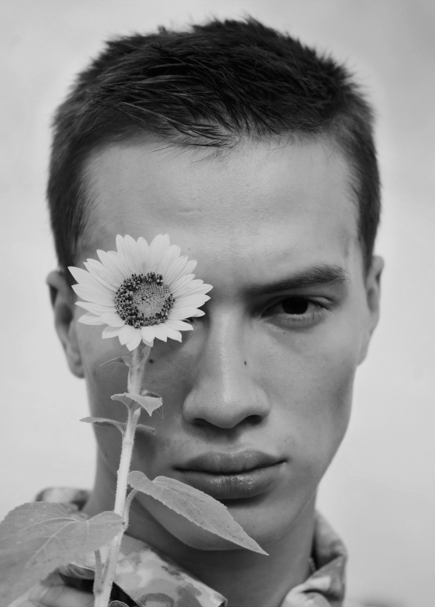 Young man holding a flower in front of one eye, looking at the camera, black and white photo.