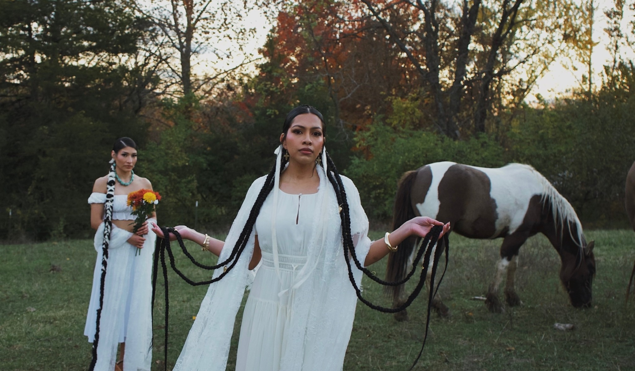 Two women in white dresses holding horse leads in a grassy field with trees, one woman in front and one in back.