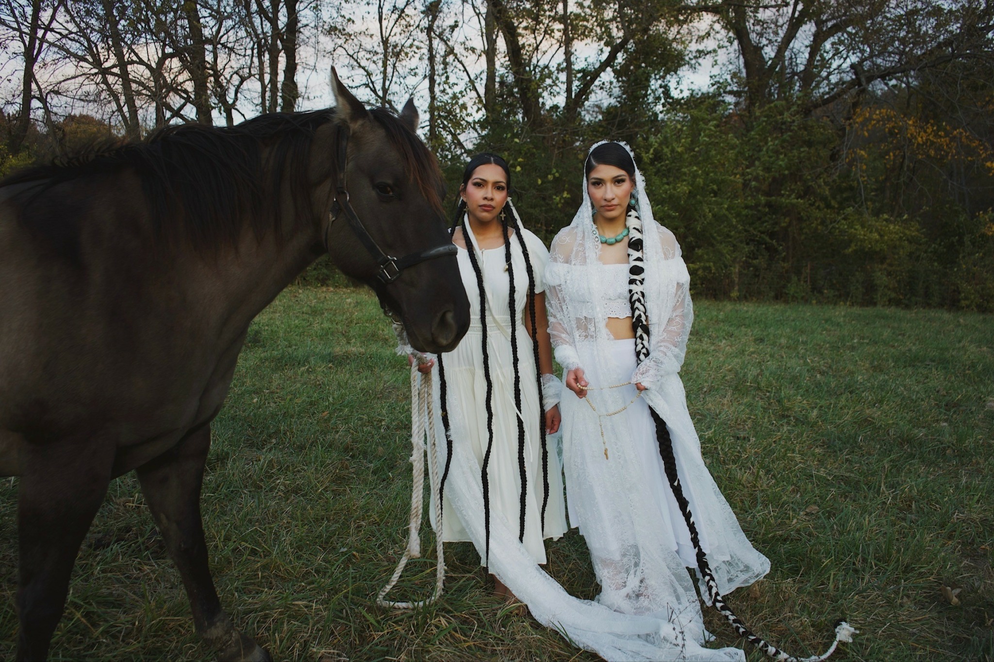 Two women in white dresses stand outdoors next to a dark horse, with trees and grass in the background.