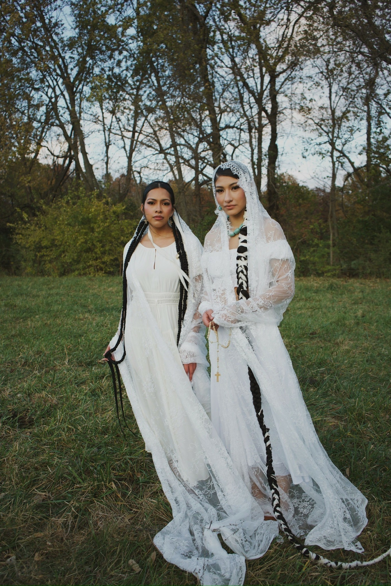 Two women in white dresses standing outdoors on grass with trees in the background.