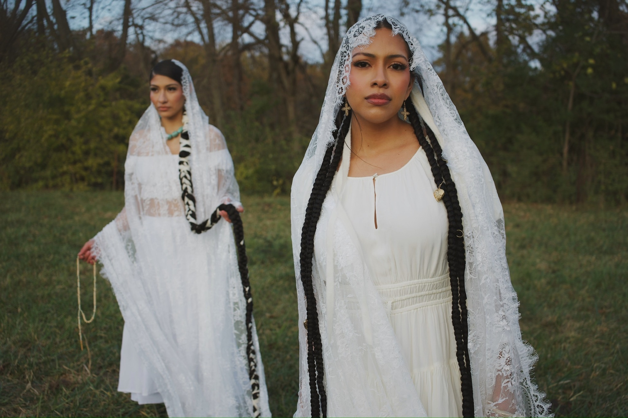 Two women in white dresses and lace veils stand outdoors with trees in the background.