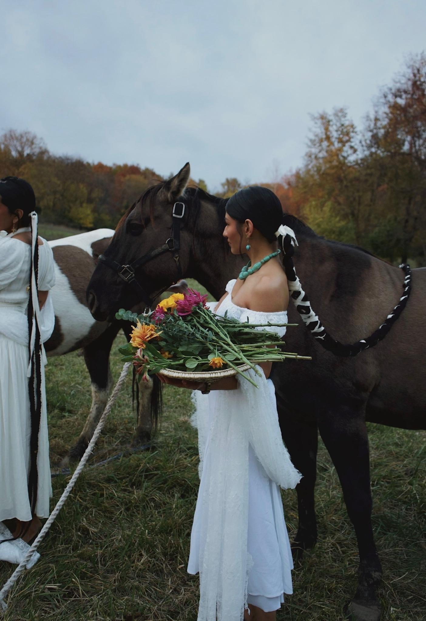 Person in white dress holding a bouquet of flowers near a horse, with another person and horse in background outdoors.