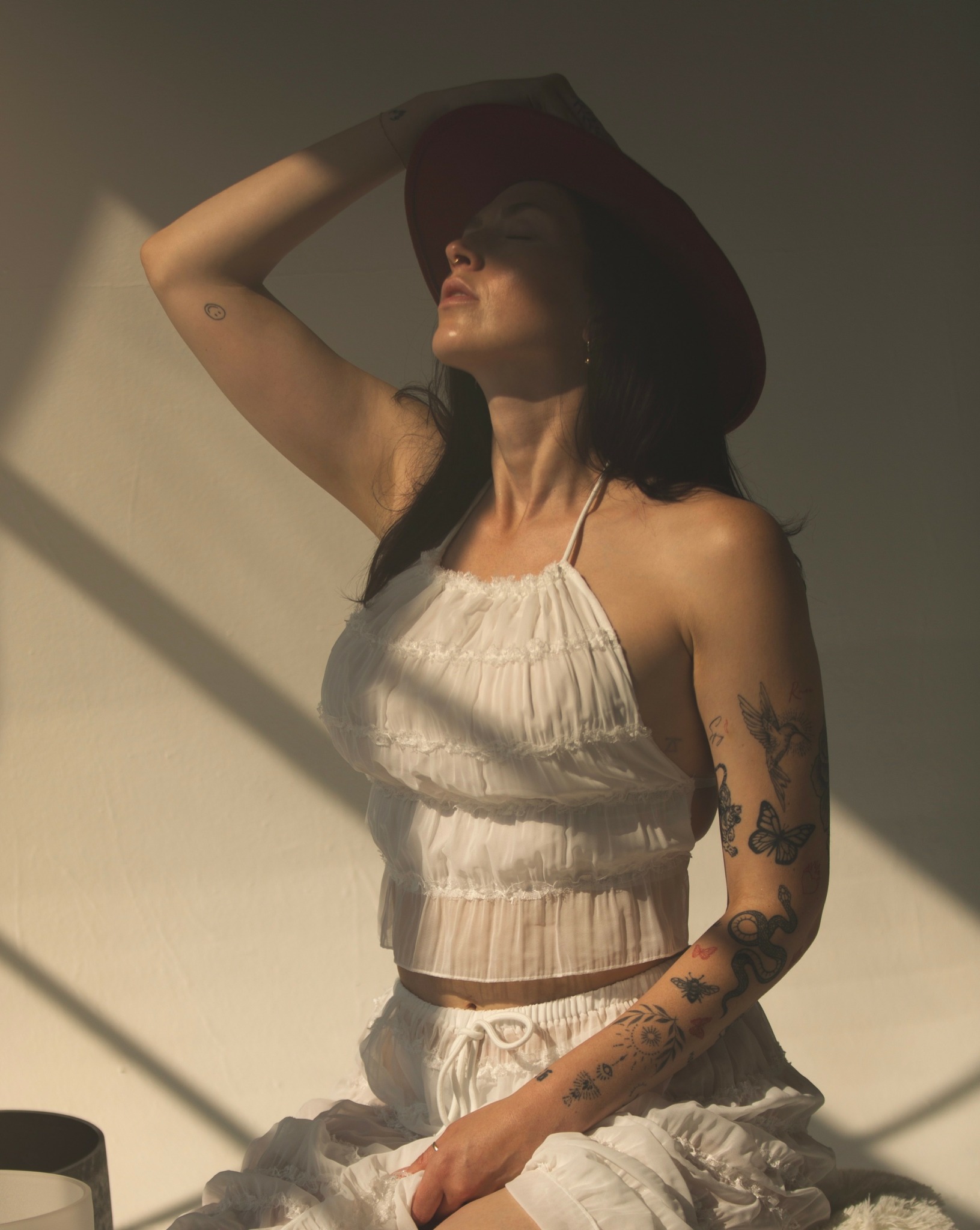 Woman wearing a large hat, white top, and pants, sitting with sunlight casting shadows on her face and body.