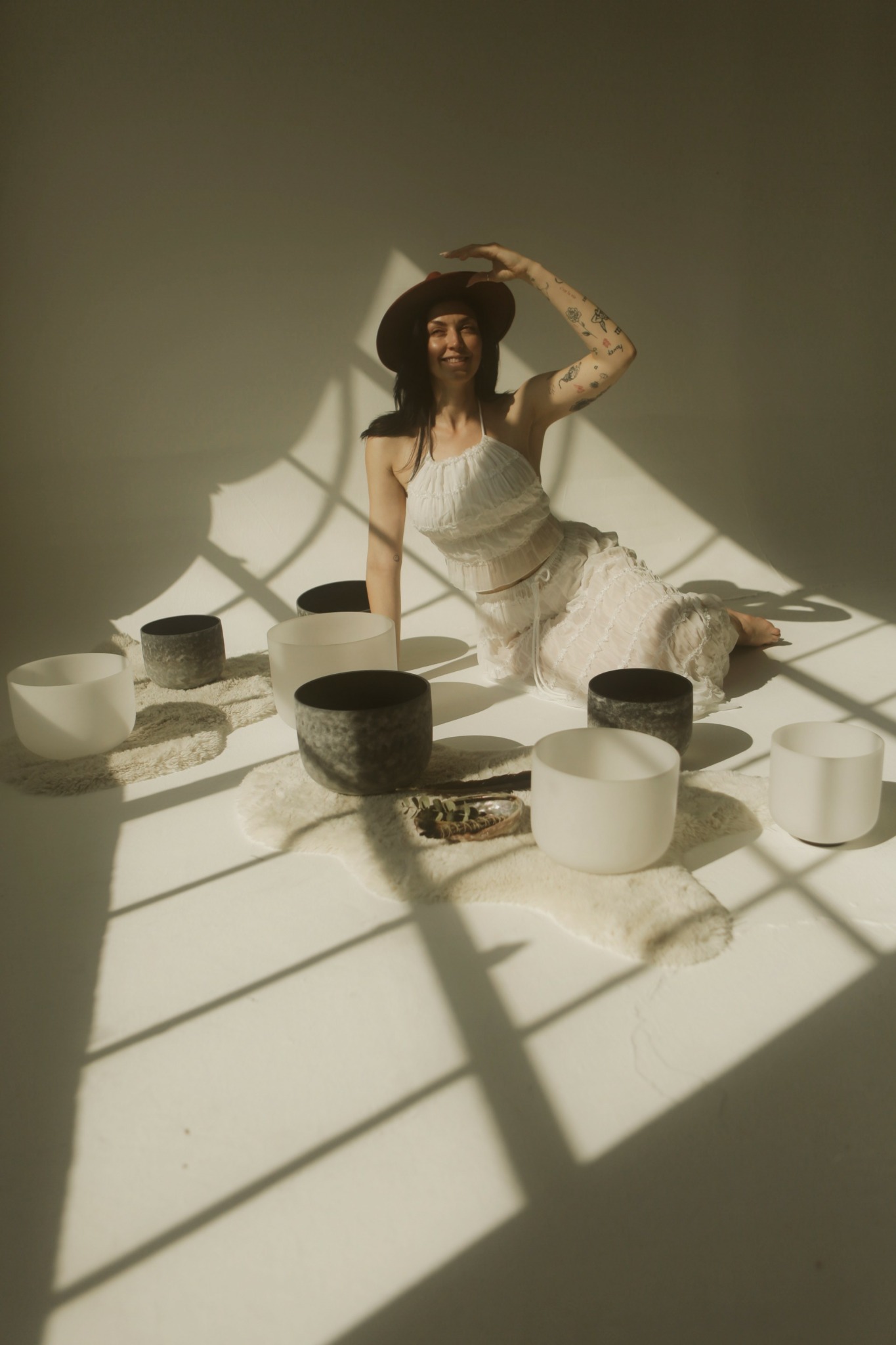 Woman in a white dress and hat sitting among black and white bowls, with shadows cast on the floor.
