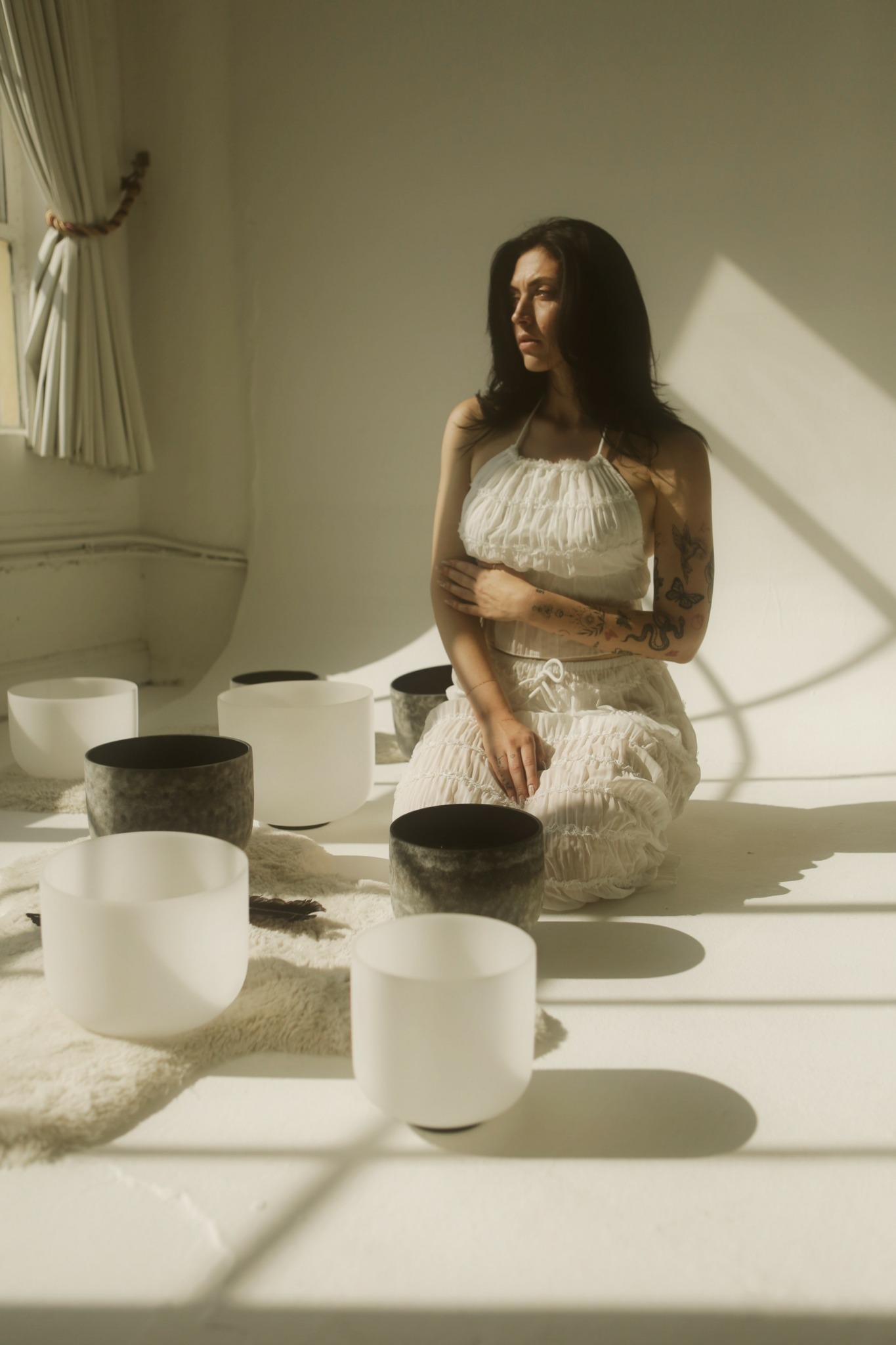 Woman with dark hair and tattoos sitting on a white surface, surrounded by white and black bowls, sunlight streaming through window.