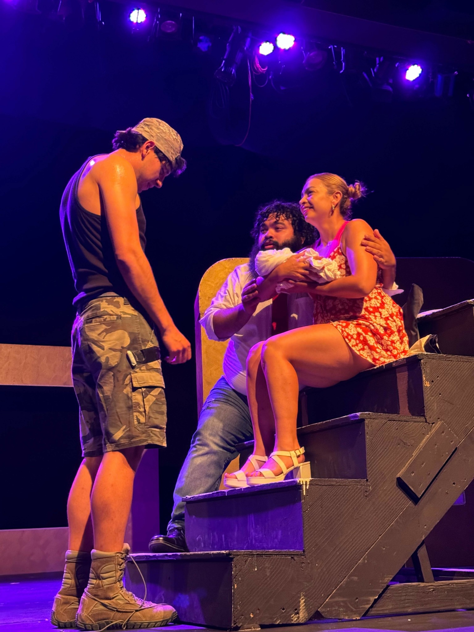 Three people on stage, one sitting on stairs, two standing, with stage lights above, in a theatrical setting.