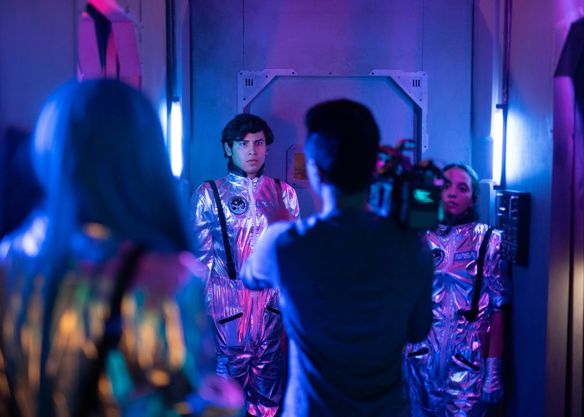People in futuristic metallic outfits in a dimly lit room with blue and purple lighting, one person being filmed.
