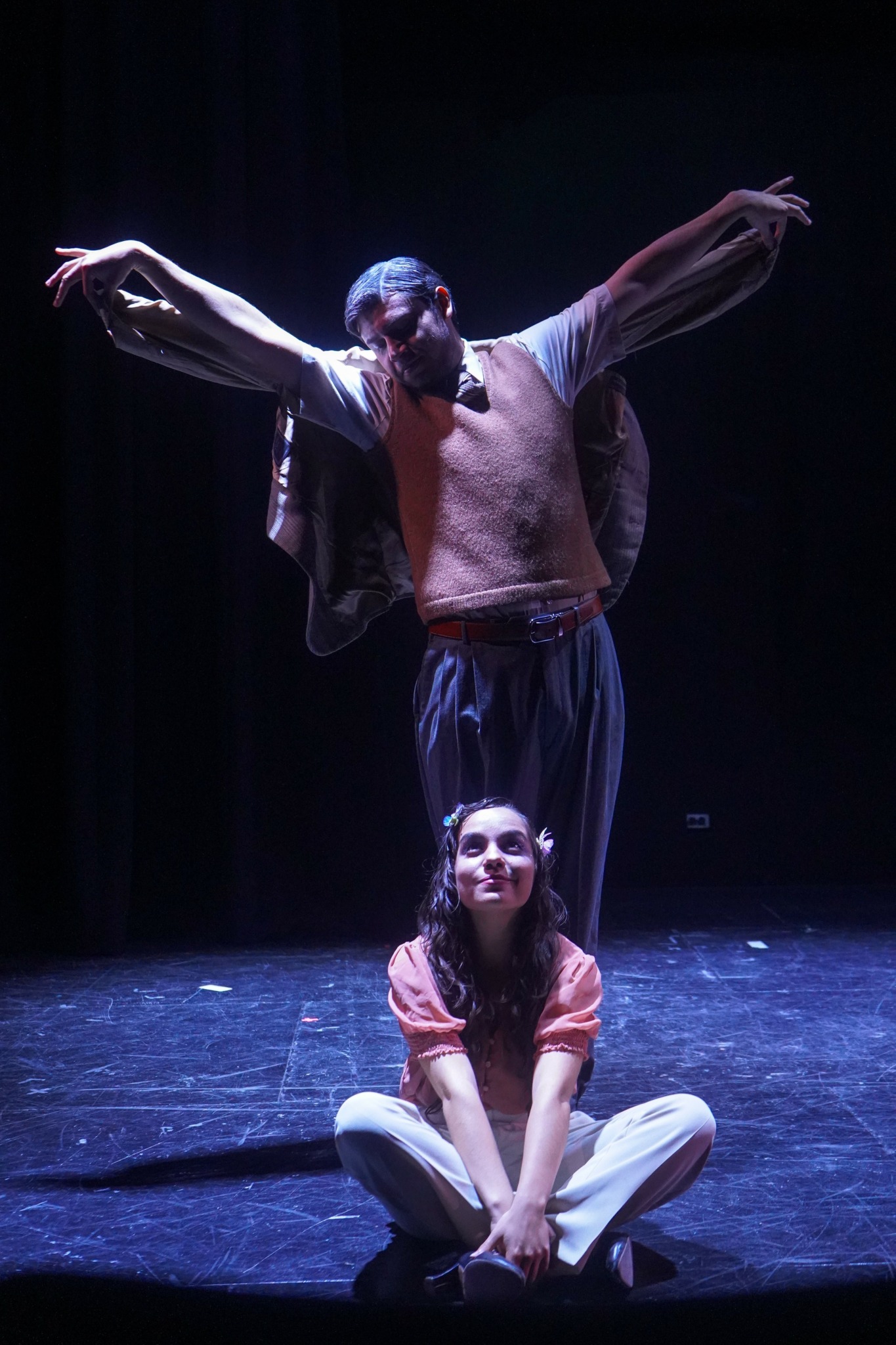 Two performers on stage, one standing with arms outstretched and the other sitting cross-legged, smiling.