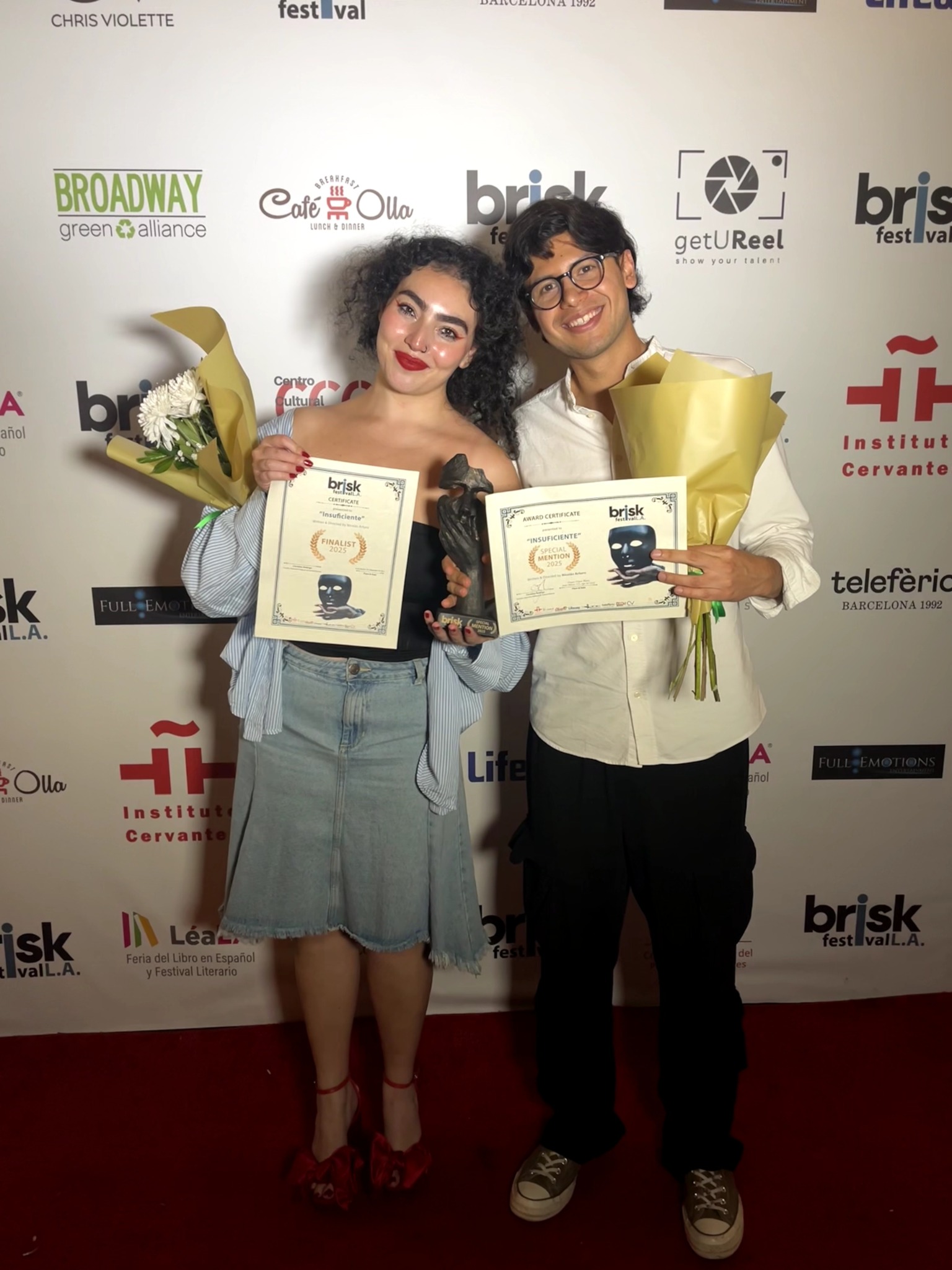 Two people holding awards and flowers, smiling in front of a backdrop with various logos.