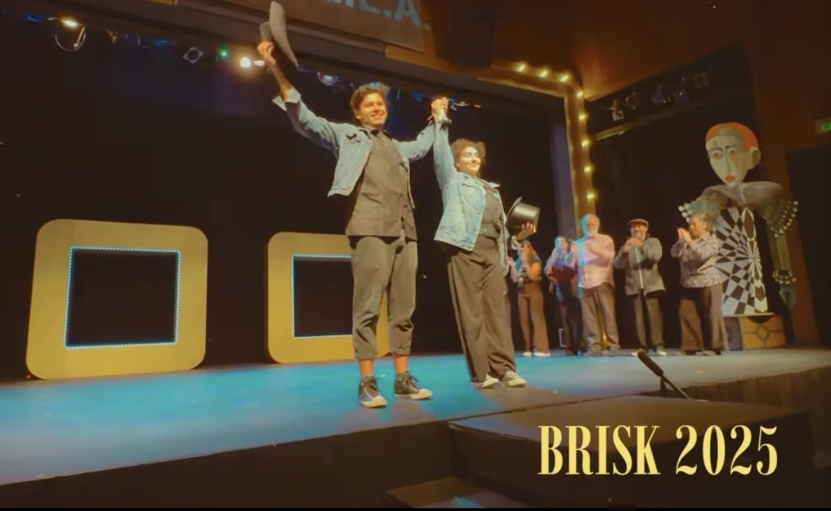 Two people stand on stage holding hands, with others in the background, stage lights, and a sign reading 'BRISK 2025'.