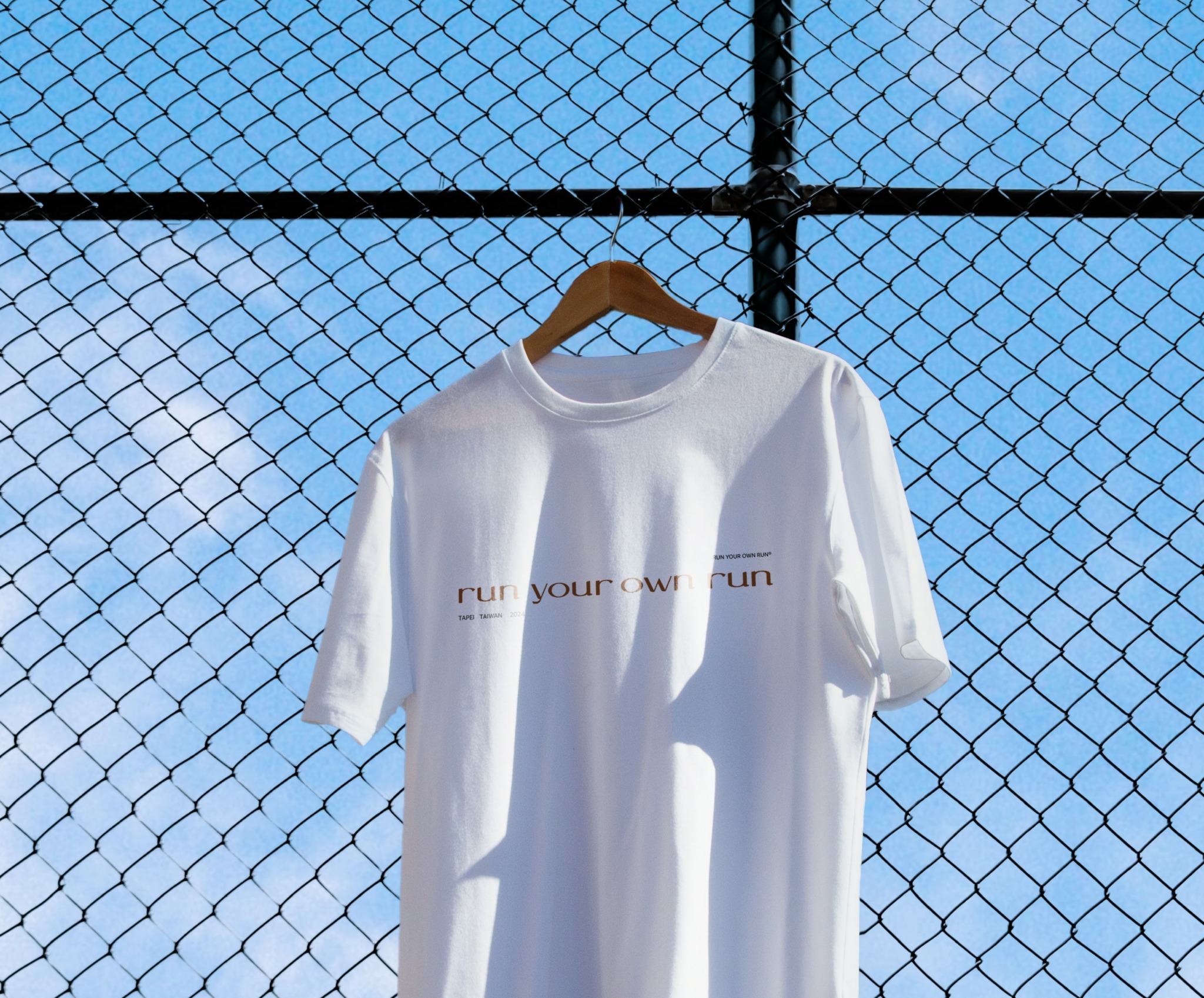 White T-shirt hanging on a hanger against a chain-link fence with a blue sky background.