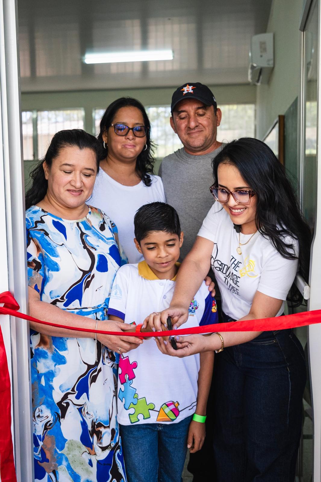 Group of five people, including children and adults, at a ribbon-cutting event indoors, smiling and holding a red ribbon.