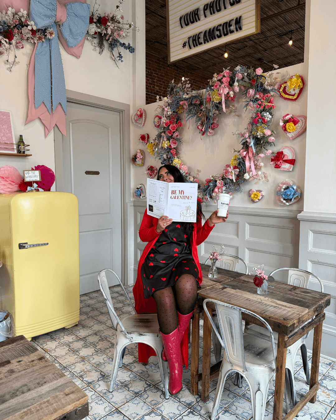 Person sitting at a table in a decorated room with floral wreaths and colorful ornaments, holding a sign. Bright yellow fridge nearby.