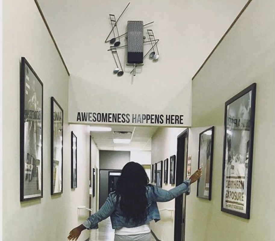 Person with long dark hair and blue shirt walking down a hallway with framed pictures, reaching out to touch a frame.