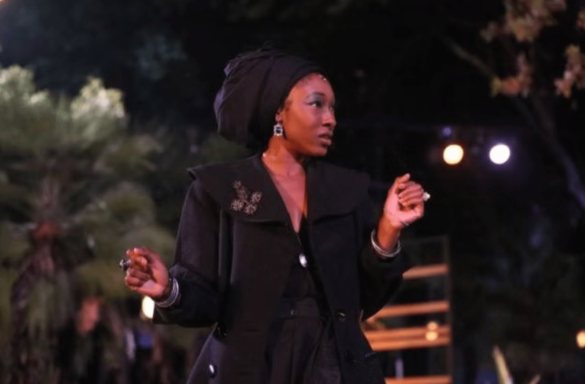 Woman wearing a headwrap and black outfit outdoors at night, with trees and lights in background.