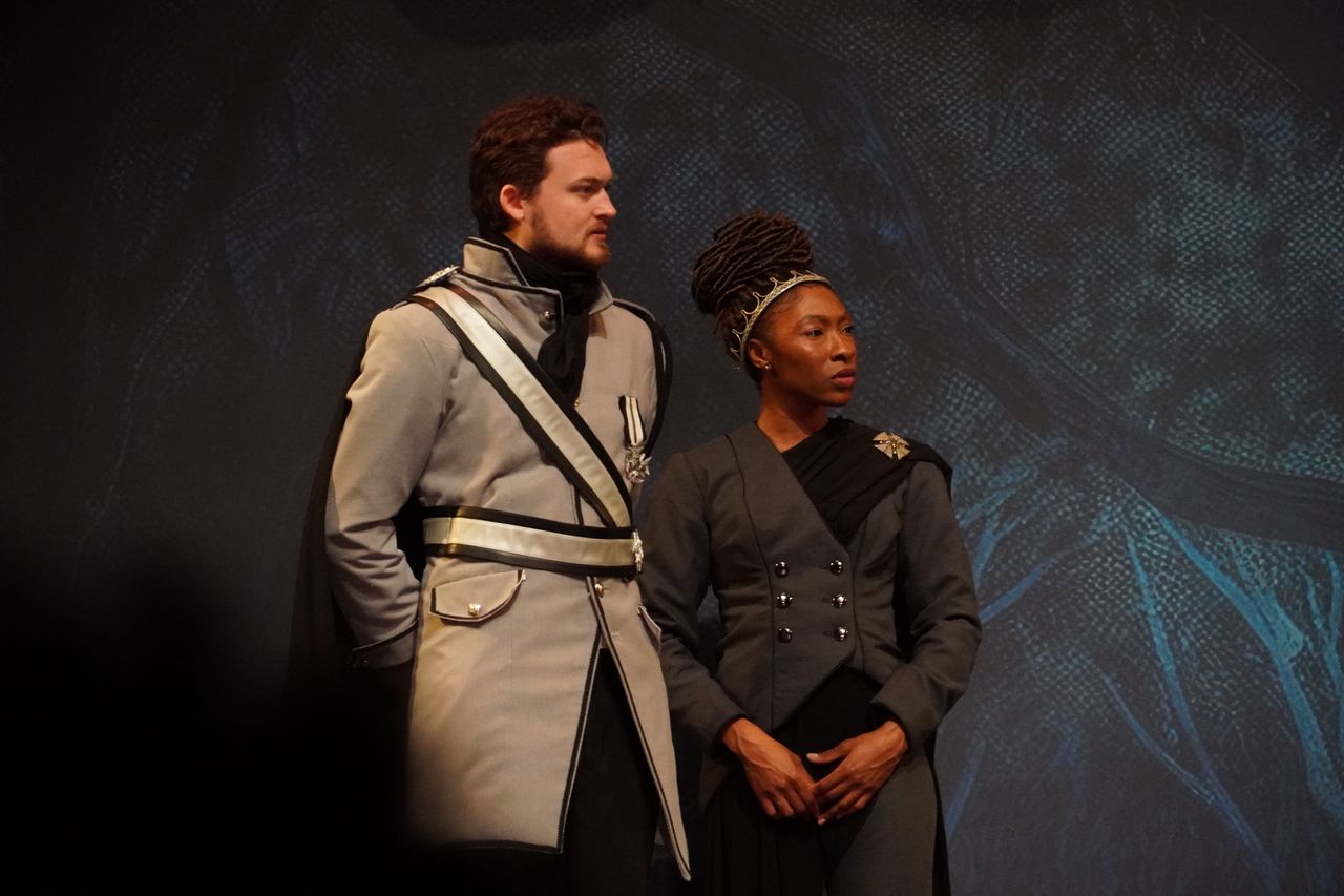 Two people stand side by side against a dark background, one in a light-colored uniform and the other in dark clothing.