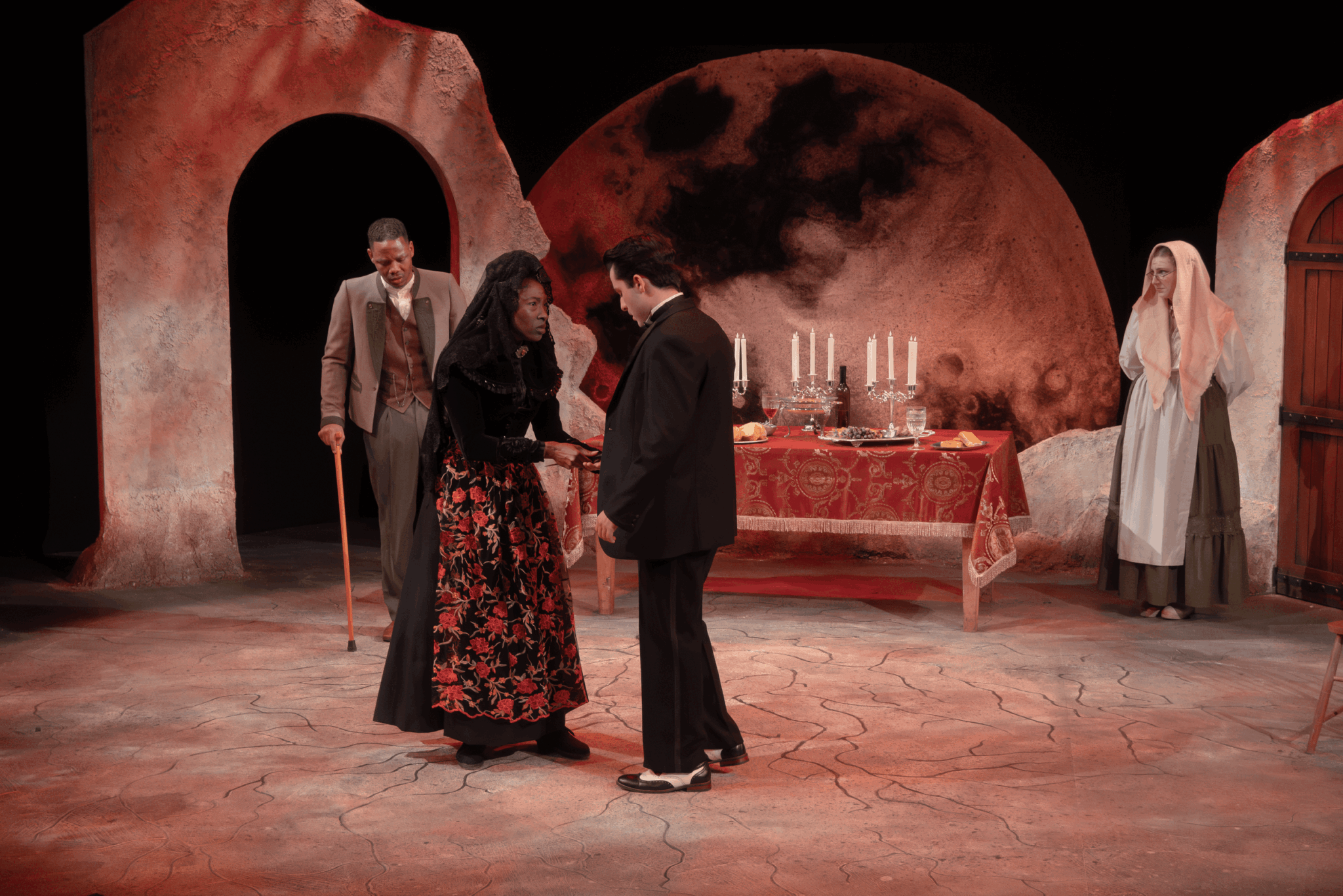 Four people on stage with a large moon backdrop, a table with candles, and an arched doorway, in a theatrical scene.
