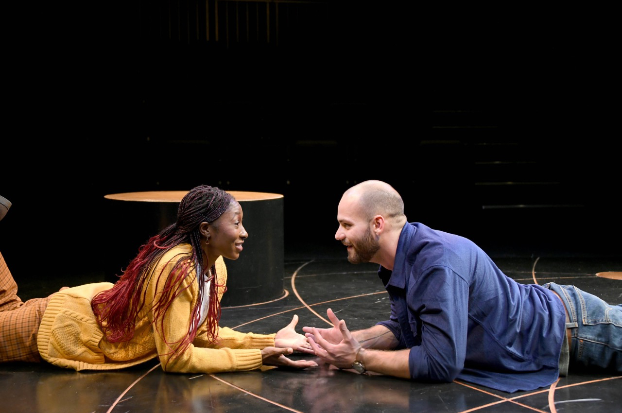 Two people lie on the floor facing each other, smiling and holding hands, with a dark background and stage lighting.