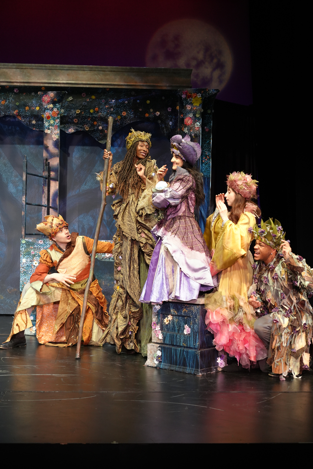 Group of actors in colorful costumes performing on stage with a moon backdrop, some standing and others seated, in a theatrical scene.