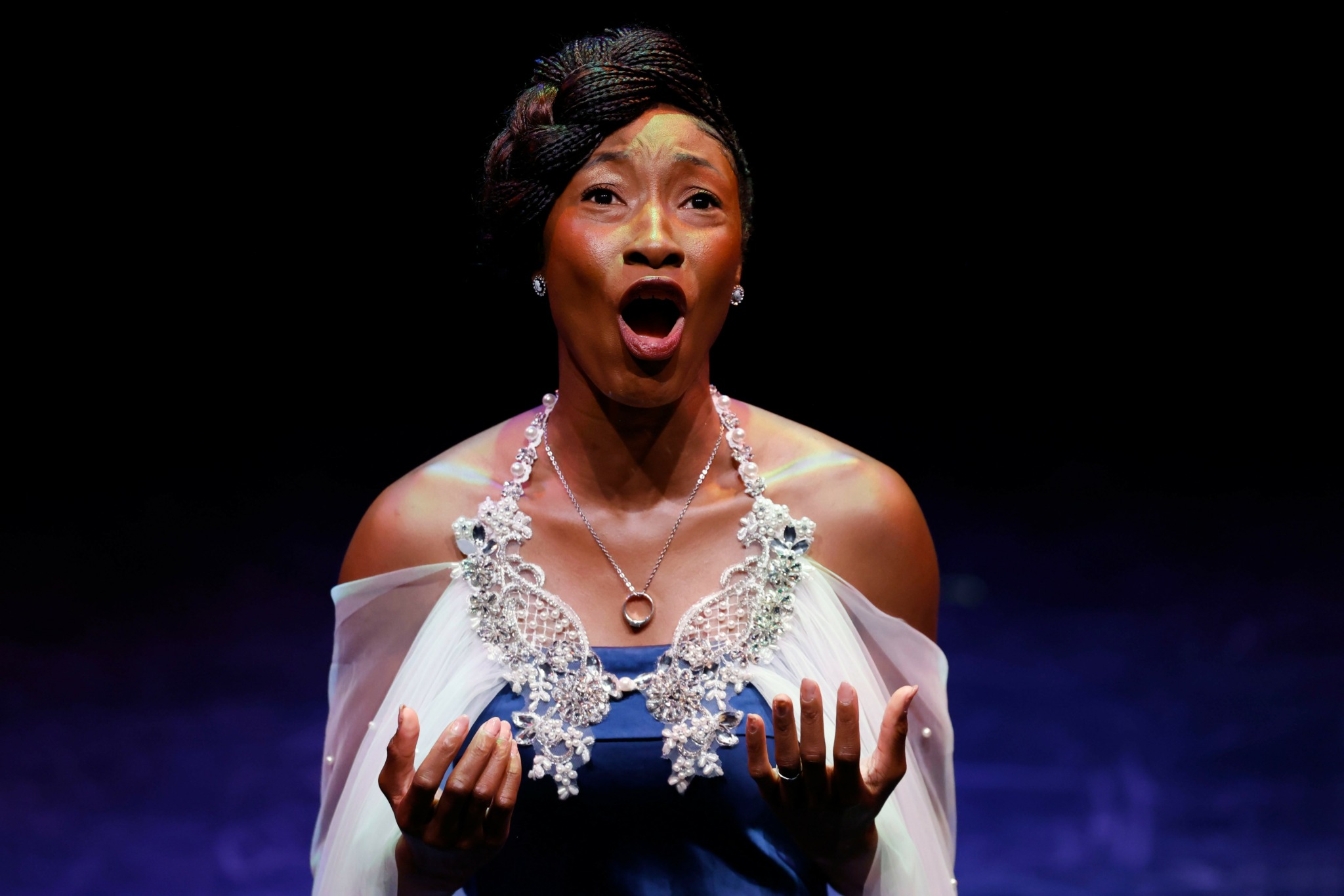 A woman with an open mouth and surprised expression, wearing a white dress with jewelry, against a dark background.