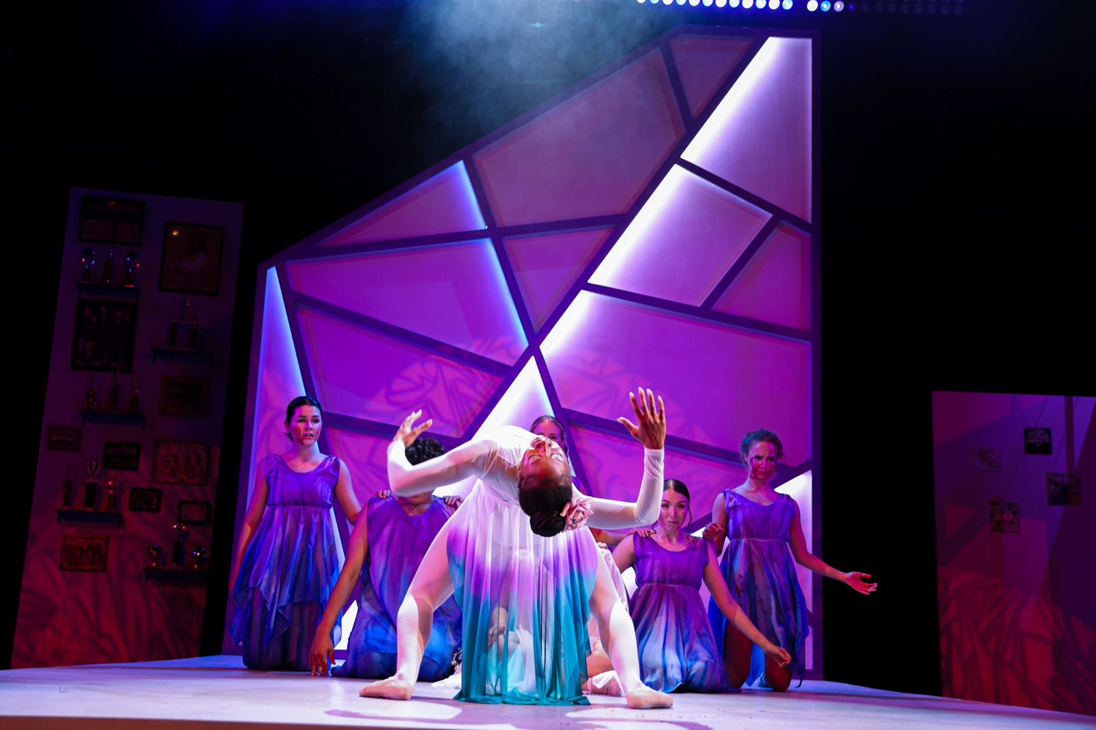 Performer in a flowing dress kneels with arms raised on stage, surrounded by four dancers in purple costumes, colorful geometric backdrop.
