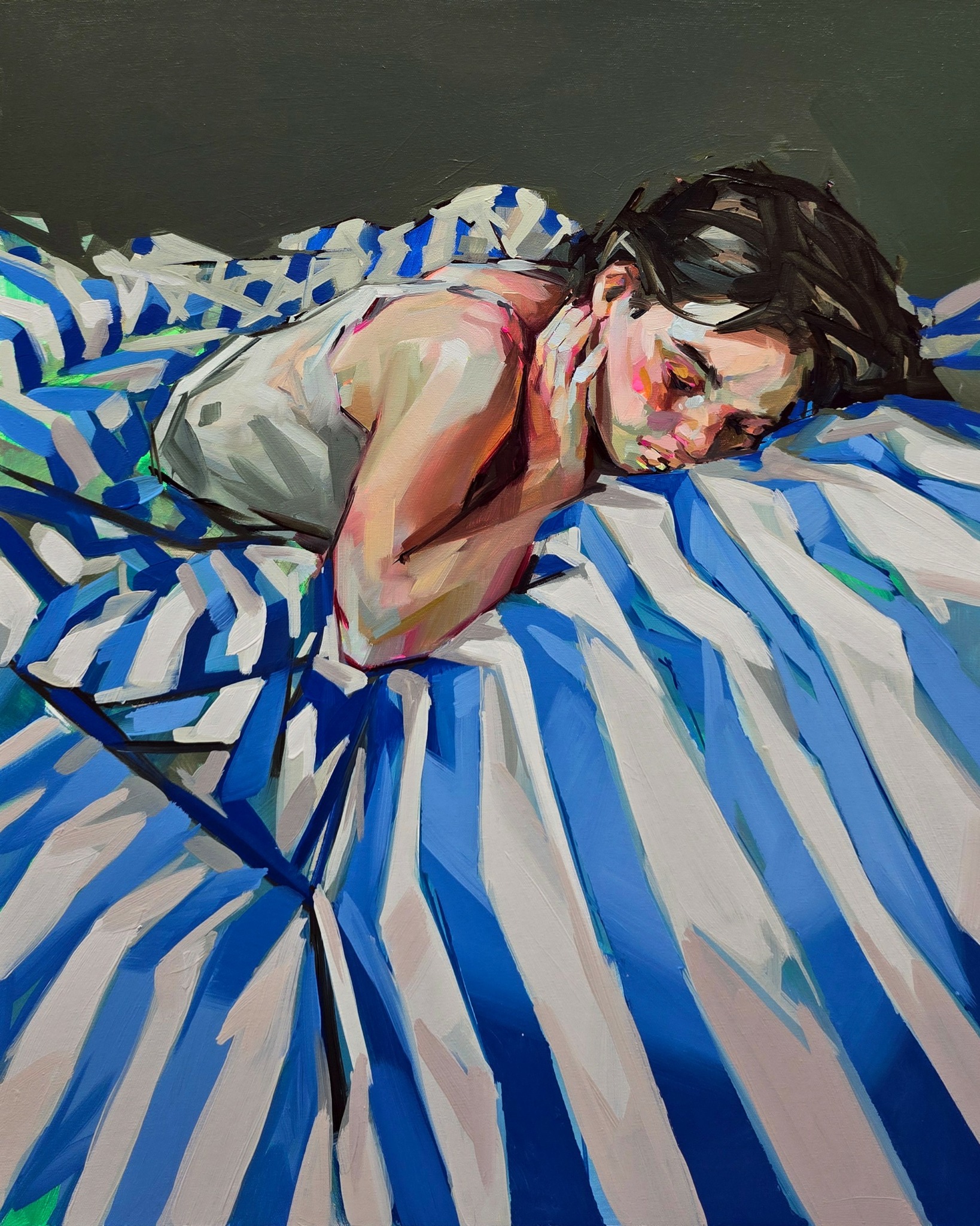 Person lying on bed with striped blue and white sheets, resting face on hand, eyes closed, dark hair, relaxed pose.