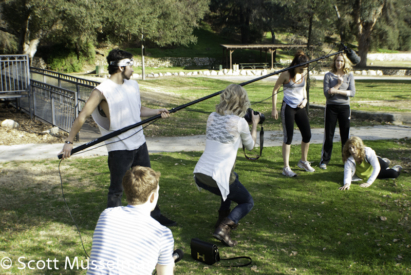 Group of people outdoors, some standing and one kneeling on grass, with trees and a fence in background.