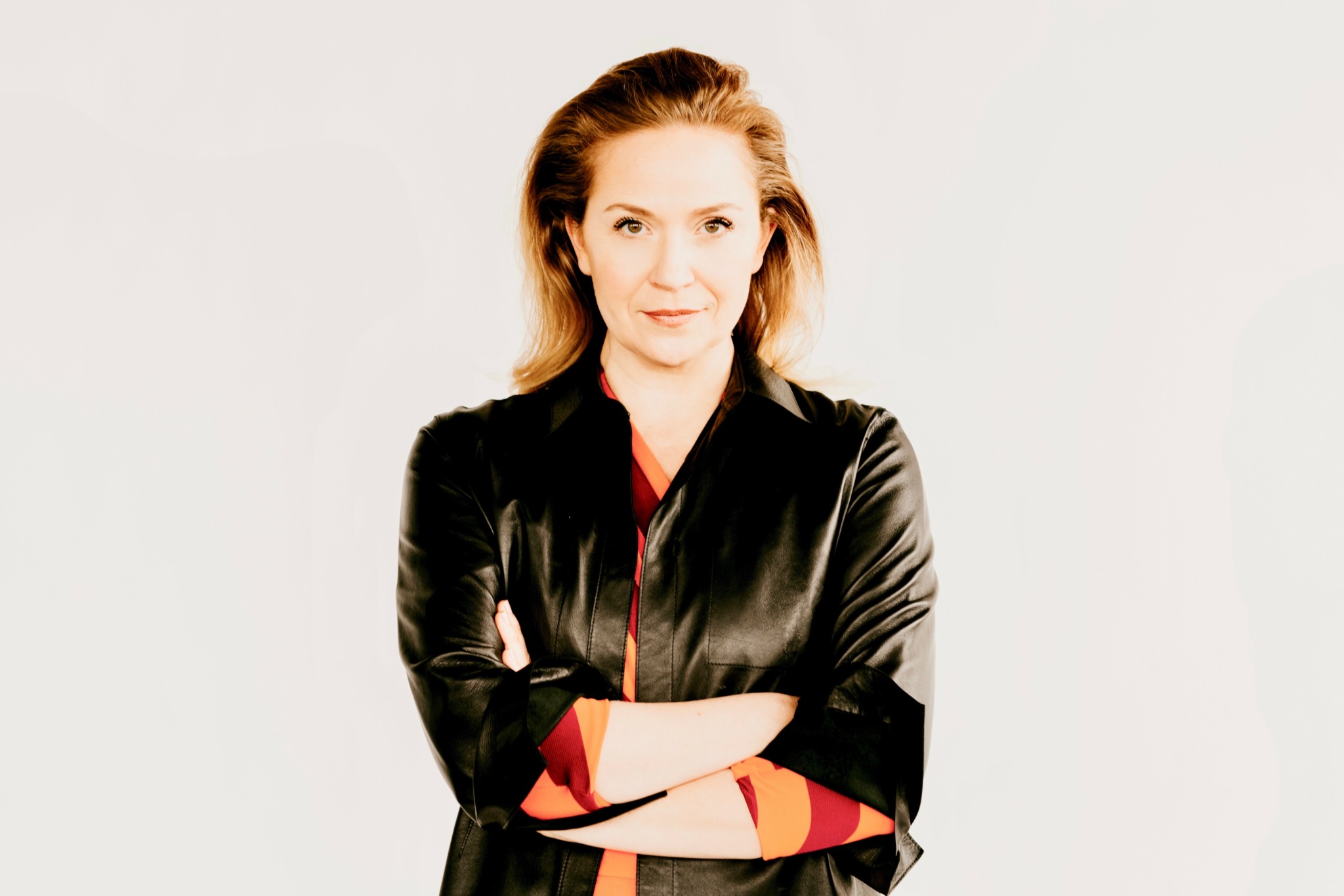 Woman with light brown hair wearing a black jacket, arms crossed, smiling, against a plain light background.