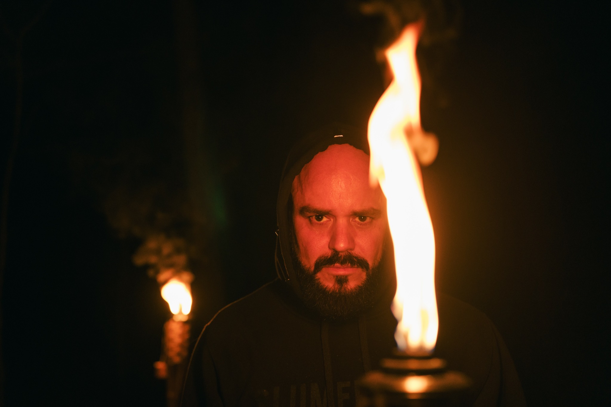 Man with long hair and beard looking at camera, with flames in the foreground and background, dark setting