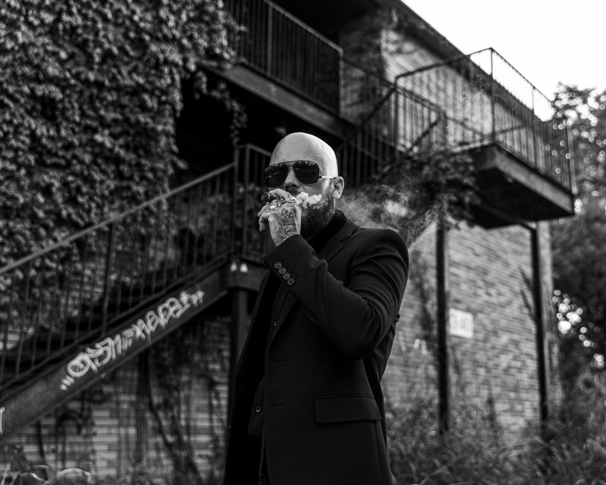 Man with sunglasses and beard in a suit, standing outdoors near a building with stairs and railing, holding a cigarette.