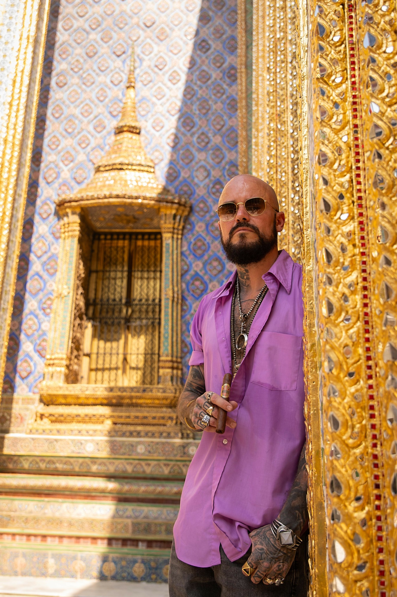 Man with sunglasses and tattoos wearing a purple shirt stands near ornate golden and blue temple architecture.