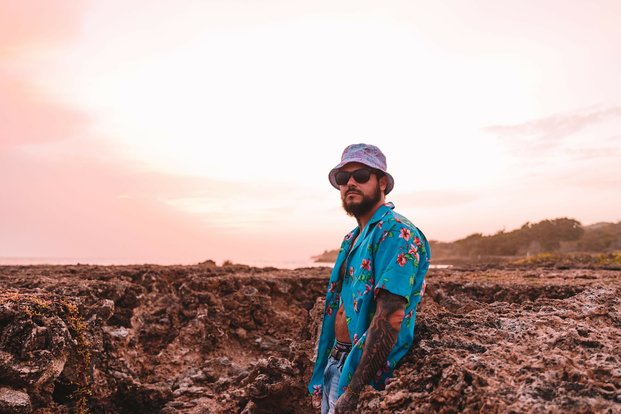 Man with a beard, sunglasses, and a hat stands outdoors during sunset, wearing a blue patterned shirt, with a rocky landscape behind him.