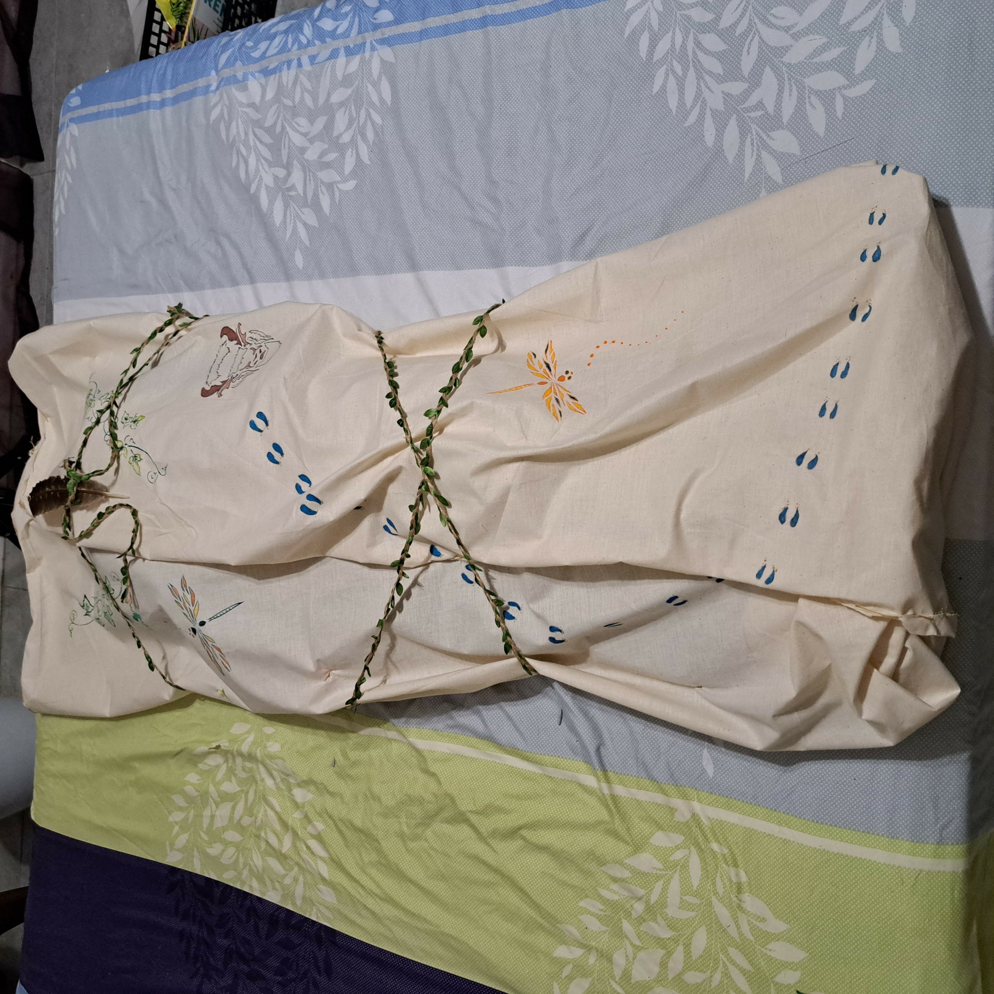 A person lying on a bed covered with a cream-colored sheet, with a vine plant draped over them, on a bed with a blue and green patterned bedsheet.