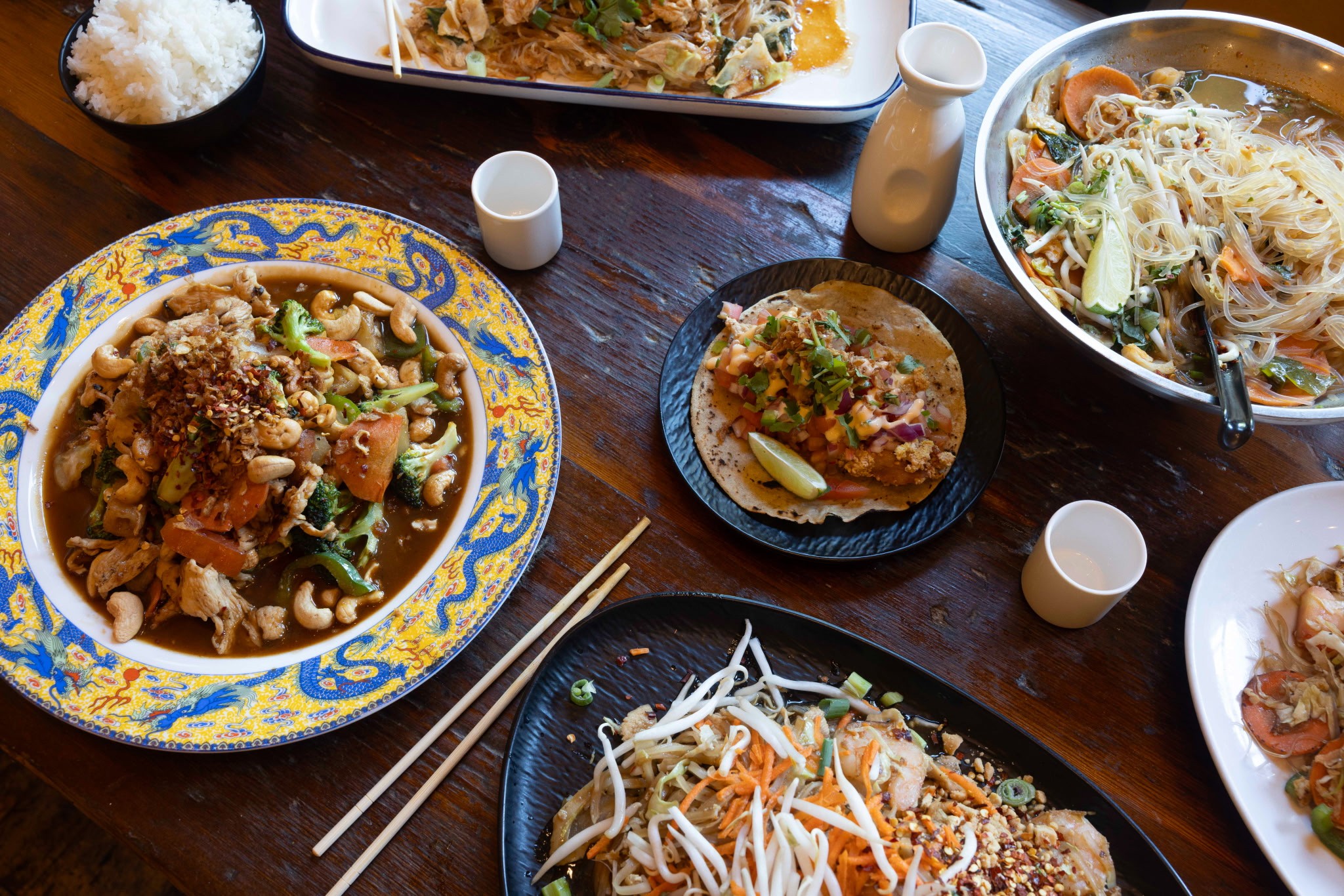 Table with various Asian dishes, bowls of rice, noodles, and sauces, chopsticks, and small cups on a wooden surface.