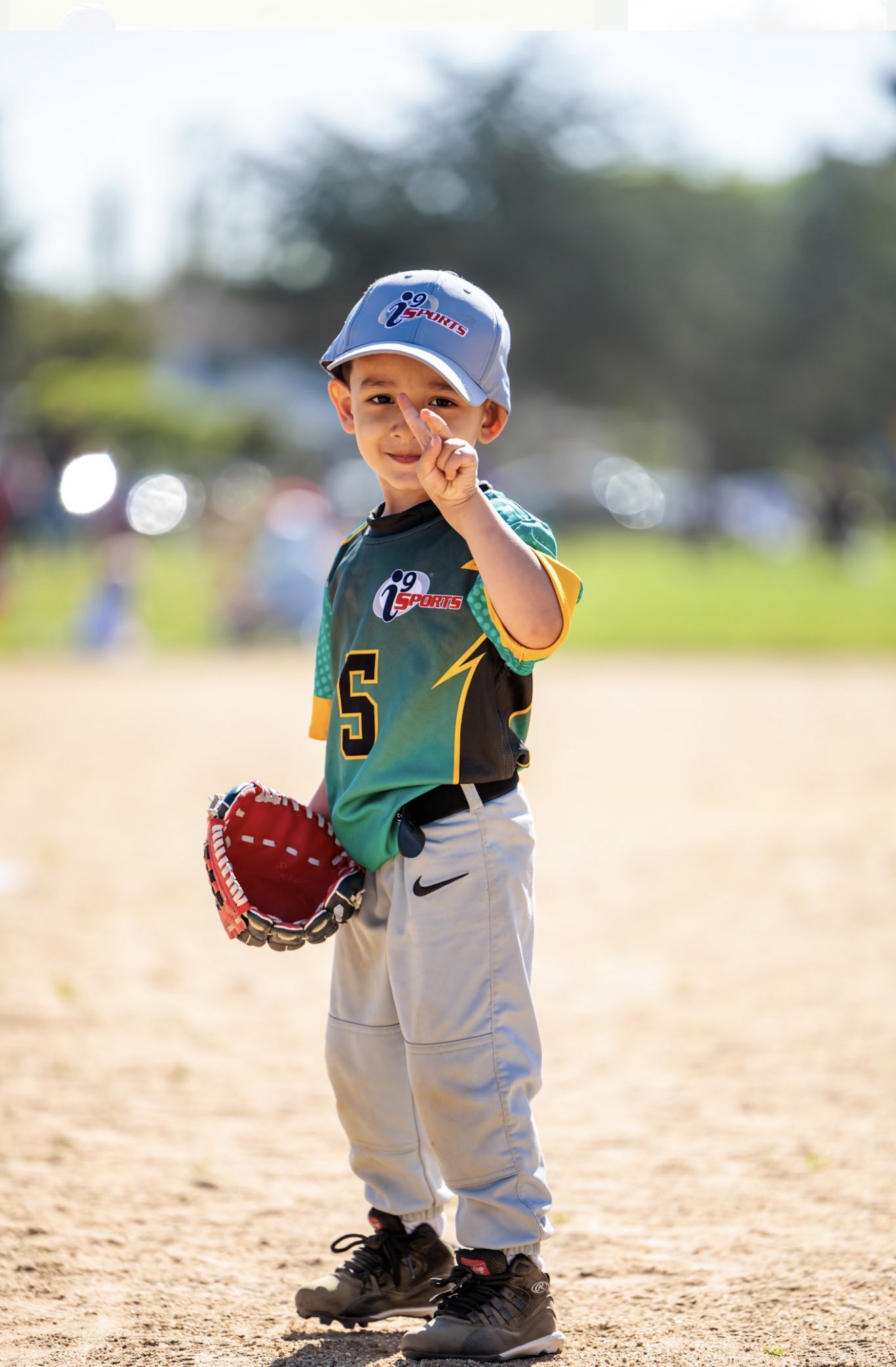 Young boy in baseball uniform holding a glove on a baseball field, smiling, with trees and sky in background.