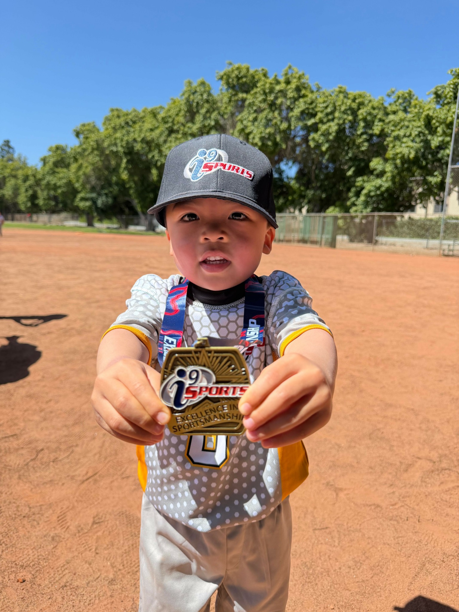 Child holding a medal on a baseball field with trees and blue sky in background.